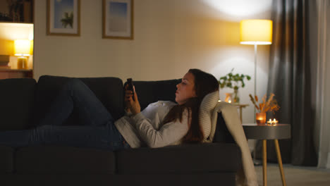 Mujer-Tumbada-En-El-Sofá-De-Casa-Viendo-Una-Película-O-Programa-O-Navegando-Por-Internet-En-Un-Teléfono-Móvil